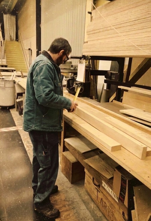 Matthew Clark in his workshop marking a board by hand before cutting, surrounded by raw lumber and shop equipment.